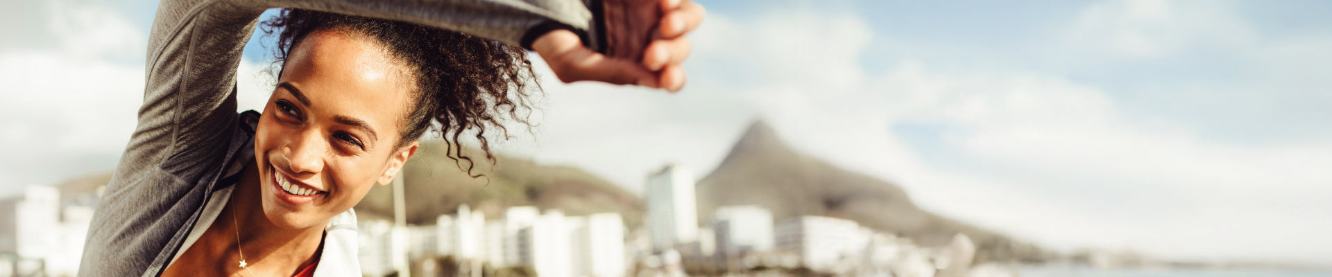 Cropped banner image of a woman with curly hair smiling and stretching outdoors, city skyline and mountain visible in the blurred background.