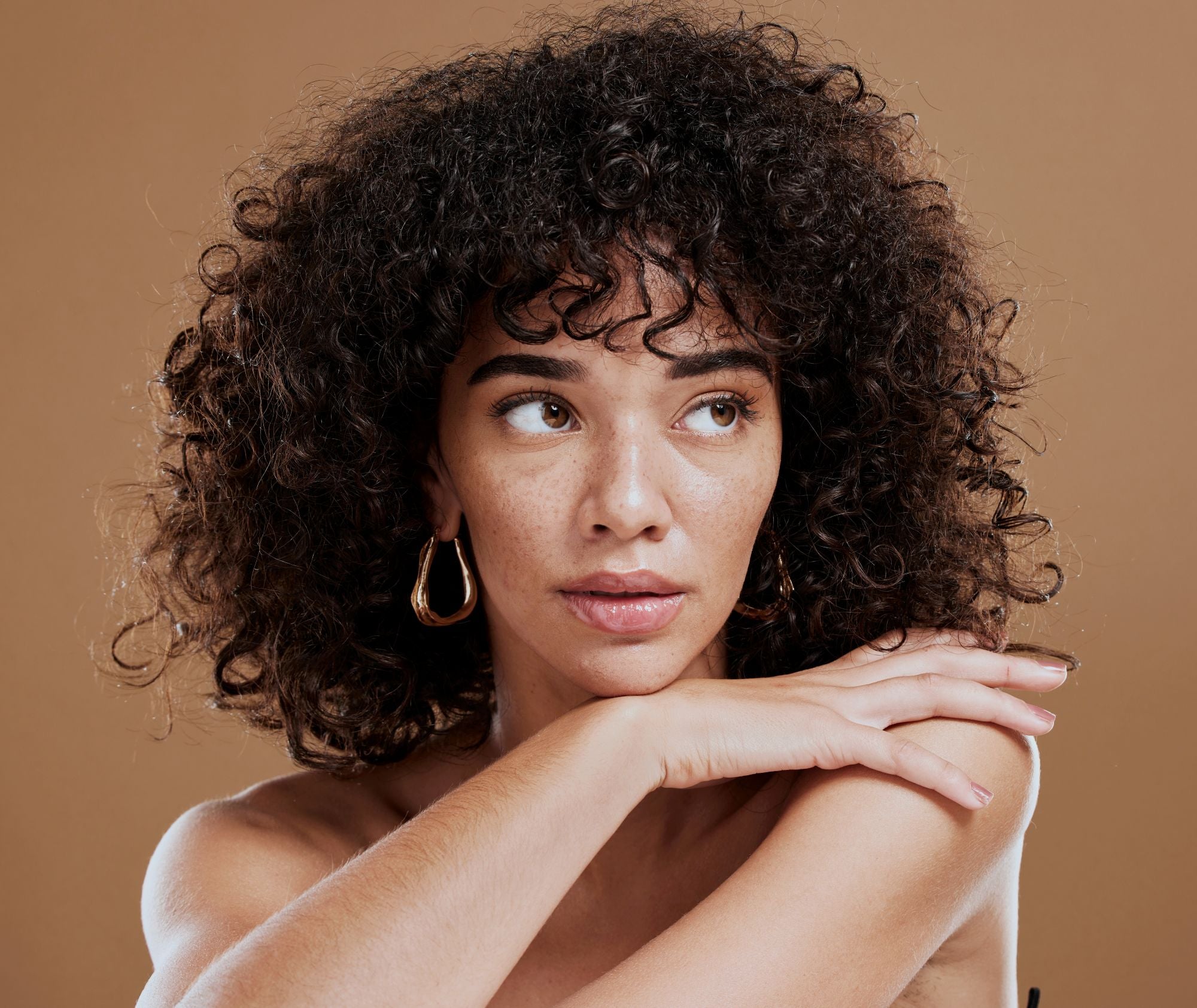 Close-up portrait of a woman with curly dark hair and gold hoop earrings, resting her chin on her crossed arms, looking off to the side against a warm brown background.