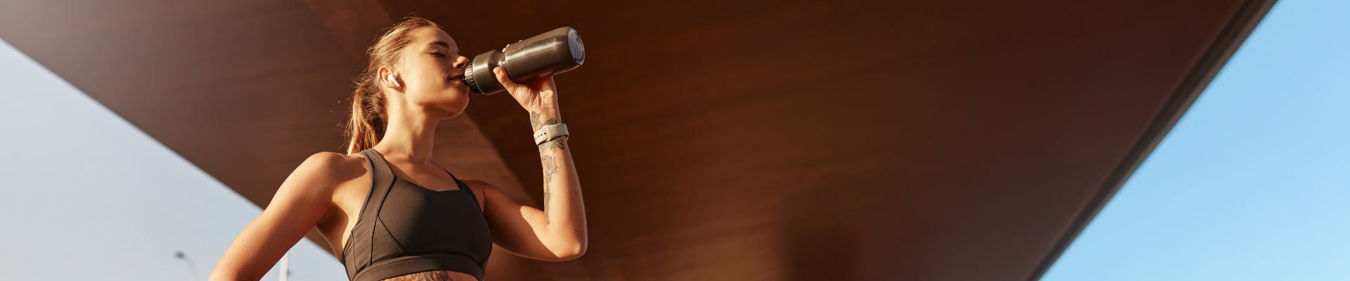 Woman in athletic wear drinking from a metal water bottle outdoors, standing beneath an angled overhang with a clear blue sky in the background.